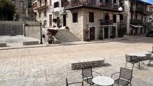 Un patio con suelo de piedra, mobiliario exterior con sillas y mesas de metal, y un edificio con varios balcones y paredes de piedra.