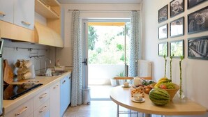 A modern kitchen with white cabinets, a dining table with fruits and a vase, and a view through a glass door to an outdoor area. - New Superb & Luminous 2 Bedroom Apartment (Kifisia)