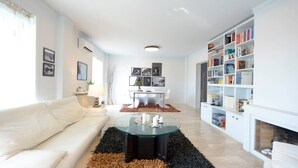 A modern living room with a white sofa, a glass coffee table, and a fireplace. There is a dining area with a white table and chairs, and a bookshelf filled with books. - New Superb & Luminous 2 Bedroom Apartment (Kifisia)