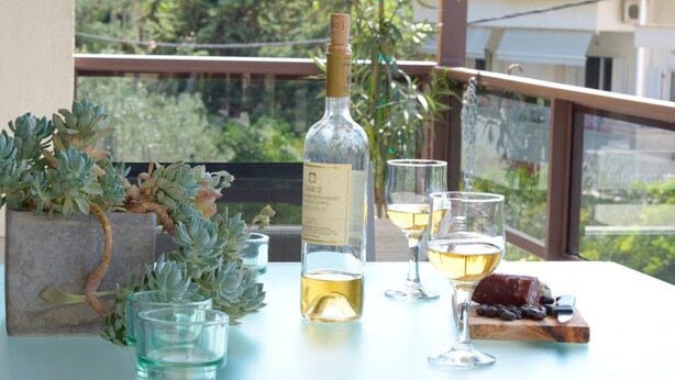 A table with a bottle of wine, glasses, and a wooden board with food on a balcony with a view of greenery and buildings. - New Superb & Luminous 2 Bedroom Apartment (Kifisia)