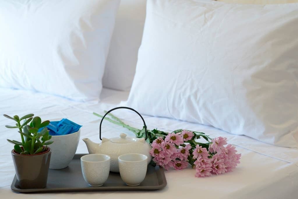 A neatly arranged tea set on a tray with a teapot, two cups, and a small potted plant on a bed with white bedding and pillows.