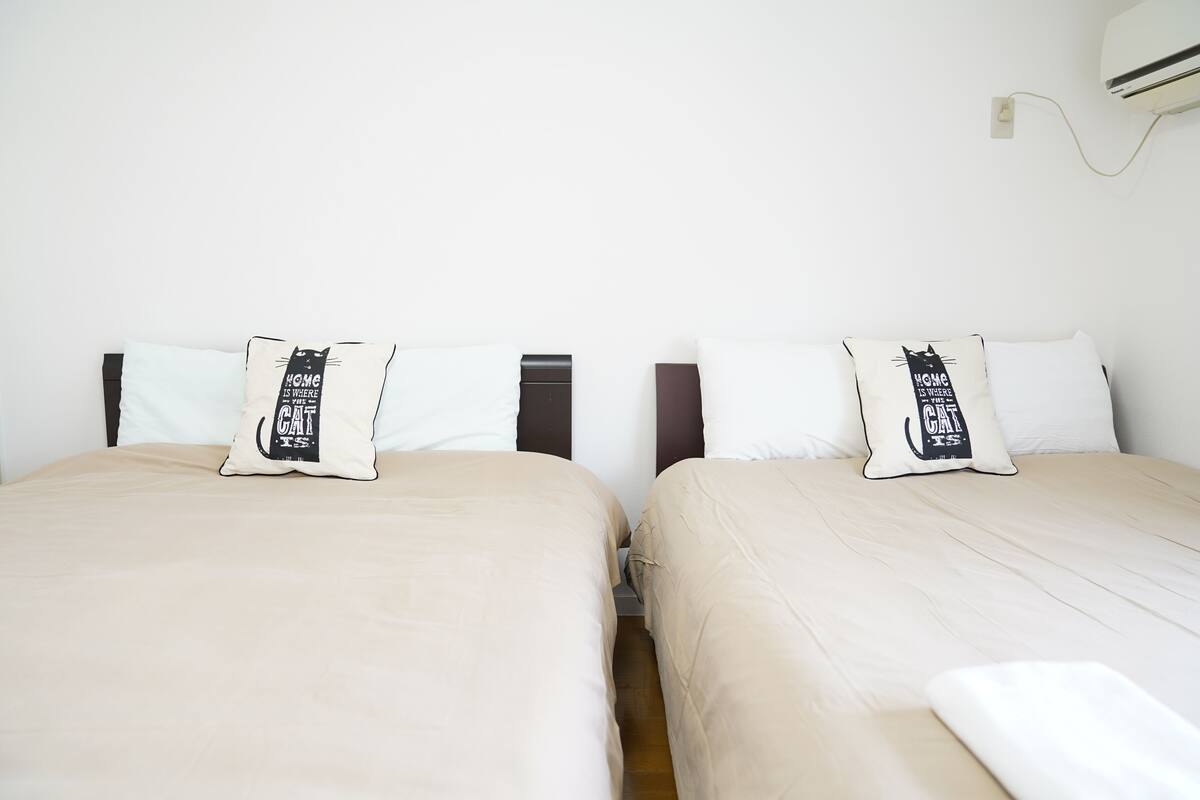 Dos camas individuales con ropa de cama color beige y almohadas a juego, una pared blanca y un aire acondicionado instalado en la pared.
