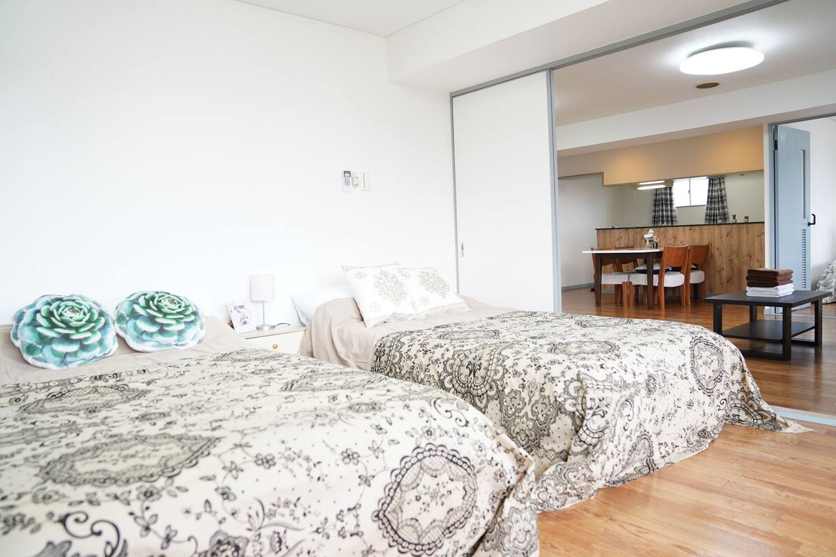 Un dormitorio con dos camas, suelo de madera, zona de comedor con mesa y sillas, y cocina al fondo.