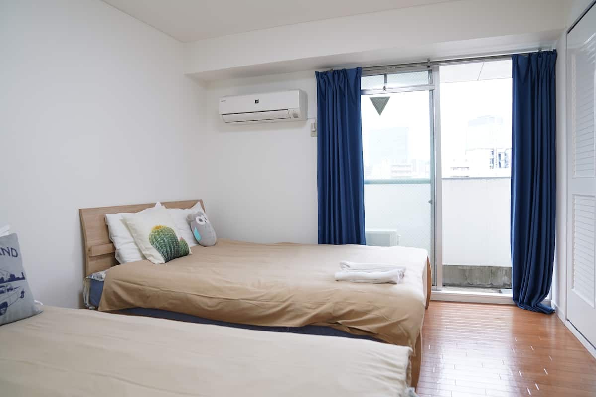 Habitación de hotel con dos camas, una ventana con cortinas azules, unidad de aire acondicionado y vistas a la ciudad.