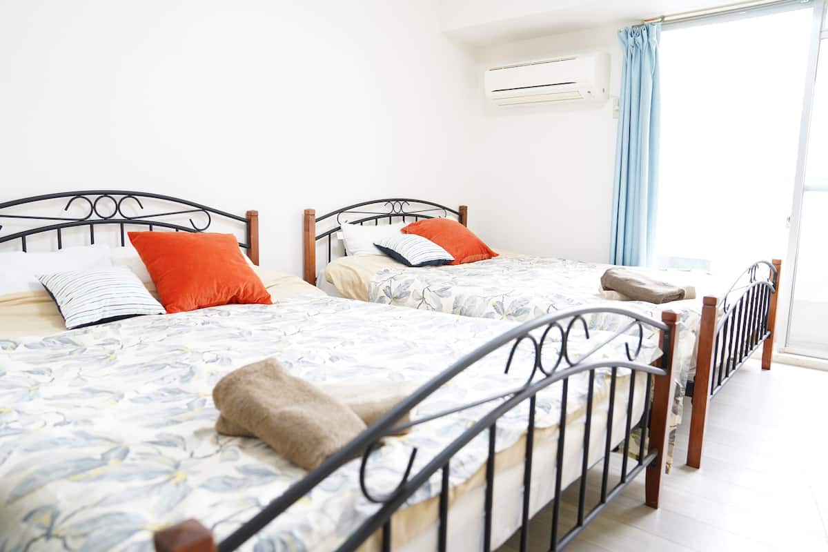 Dos camas individuales con estructura de metal y ropa de cama estampada, una ventana con cortina y un equipo de aire acondicionado en la pared.