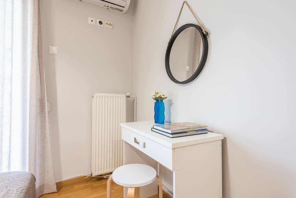 A small, neatly organized bedroom with a white desk, a round mirror, a radiator, and a window with curtains.