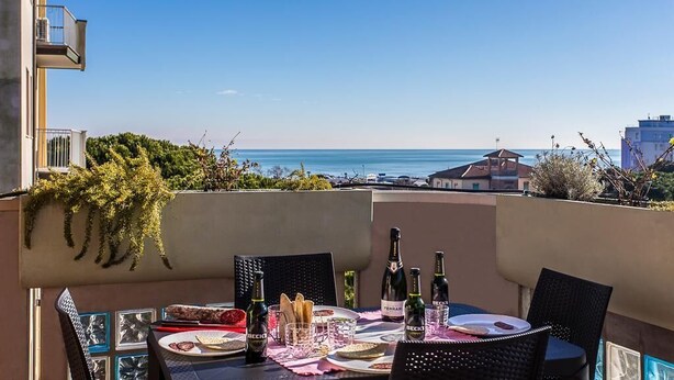 Mesa en balcón con platos, vasos y botellas, con vista al mar.
