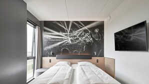 Habitació d'hotel moderna amb un llit gran, un panell de fusta al capçal, un finestra gran i un televisor fixat a la paret.