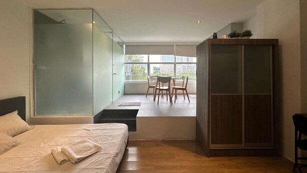 Una habitación de hotel moderna con cama, zona de estar con mesa y sillas, armario y vistas a la ciudad a través de amplias ventanas.
