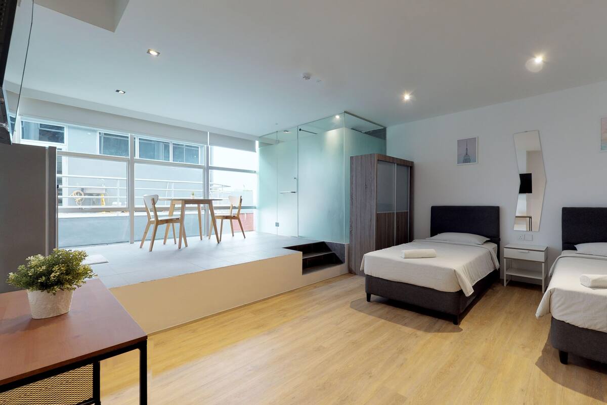 Una moderna camera d'hotel con due letti, una zona pranzo con tavolo e sedie, e un'ampia finestra che offre vista all'esterno.