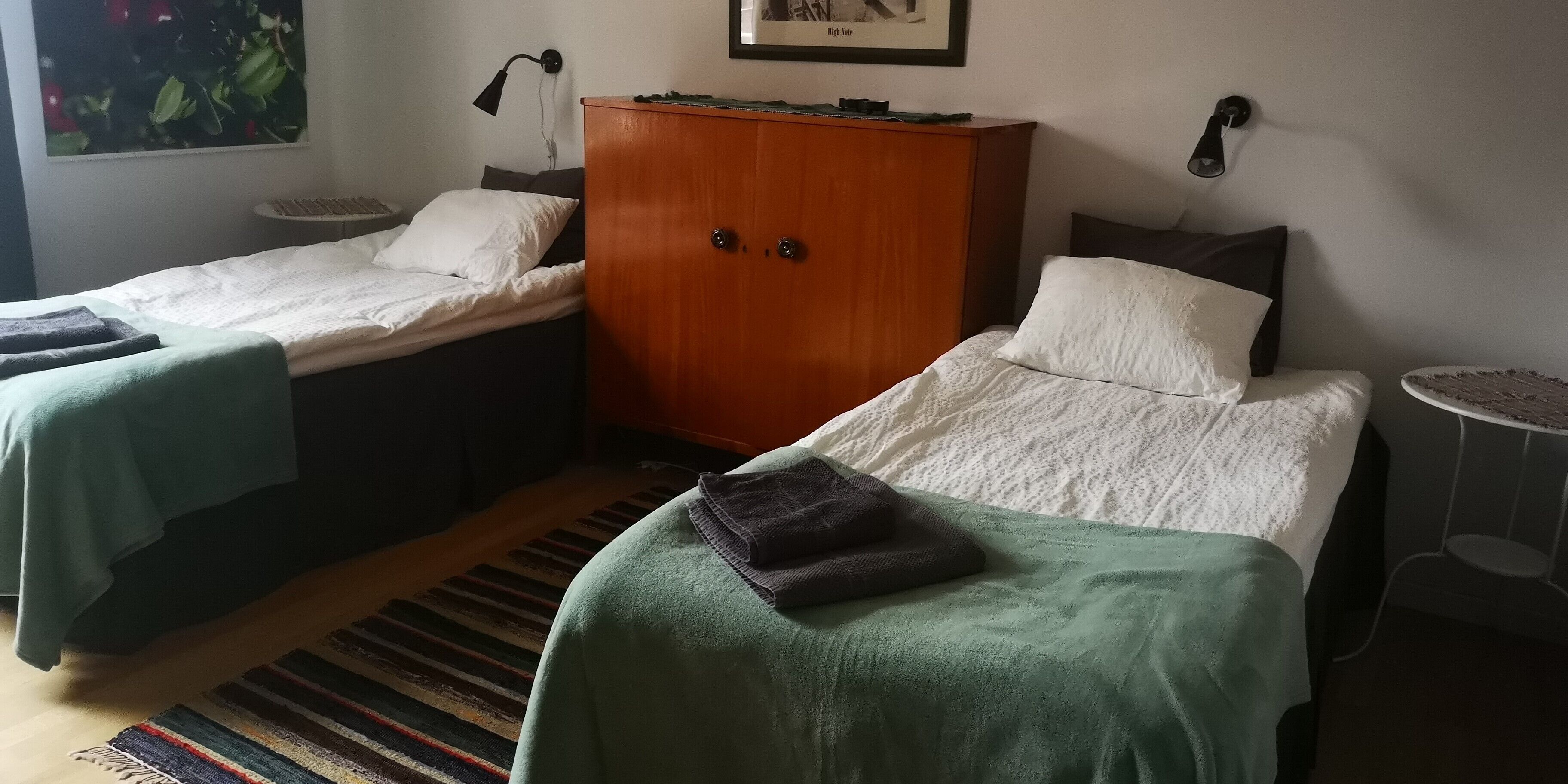 Ett hotellrum med två separata sängar, en garderob i trä, ett litet runt bord och en randig matta.