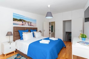 Um quarto moderno com cama, mesa de cabeceira e uma grande pintura de uma paisagem praiana.