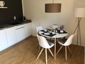 Une cuisine moderne avec des meubles blancs, une table de salle à manger ronde accompagnée de deux chaises blanches, une machine à café et un luminaire suspendu.