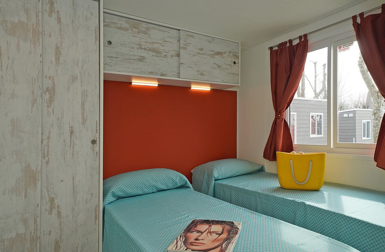 A room with two beds, a wooden wardrobe, a window with curtains, and a yellow bag on the windowsill.