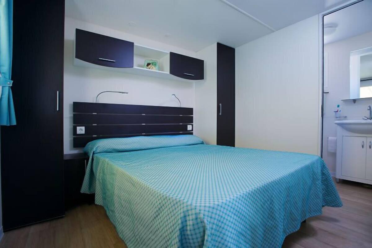 A bedroom with a bed, a built-in wardrobe, a shelf with items, and a bathroom with a sink and mirror.