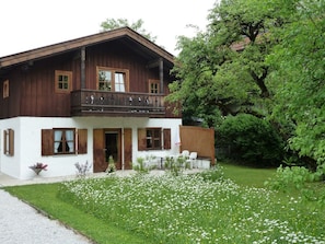 A two-story wooden house with a balcony, surrounded by a garden with white flowers.