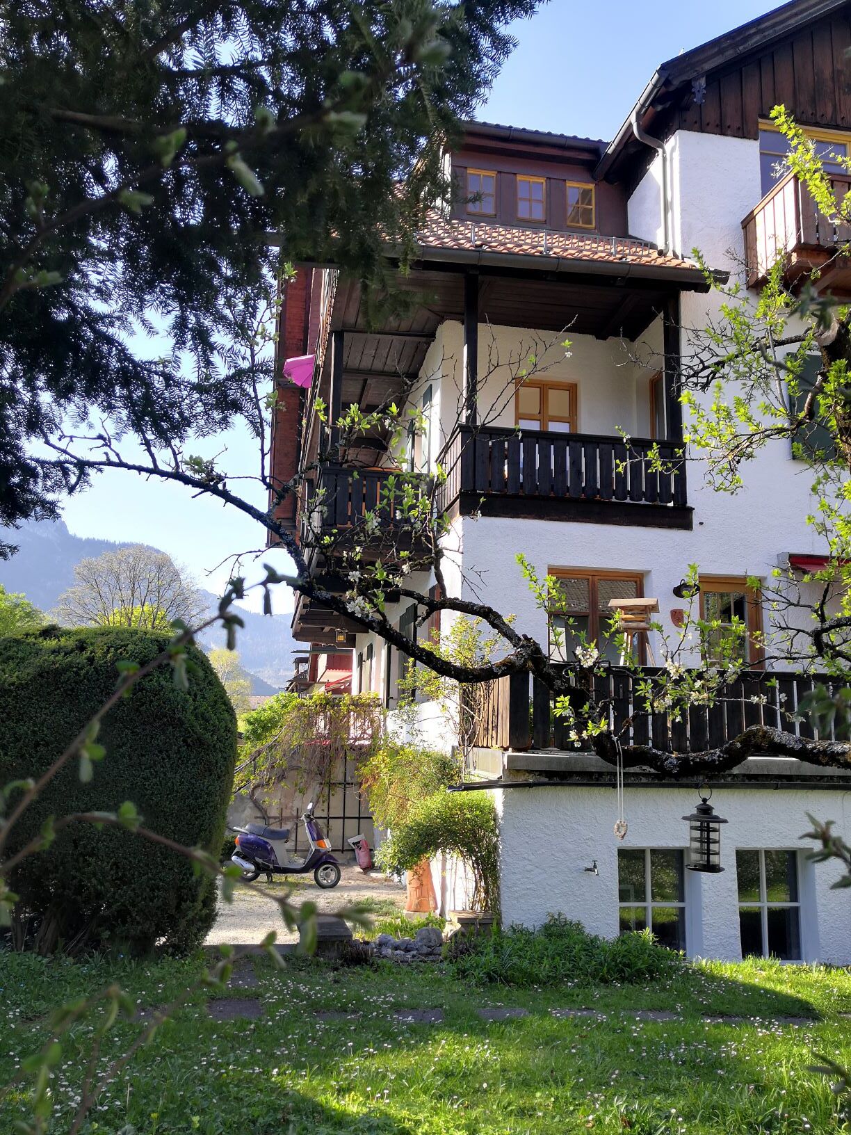 A traditional chalet-style building with a balcony, surrounded by lush greenery and a clear sky.