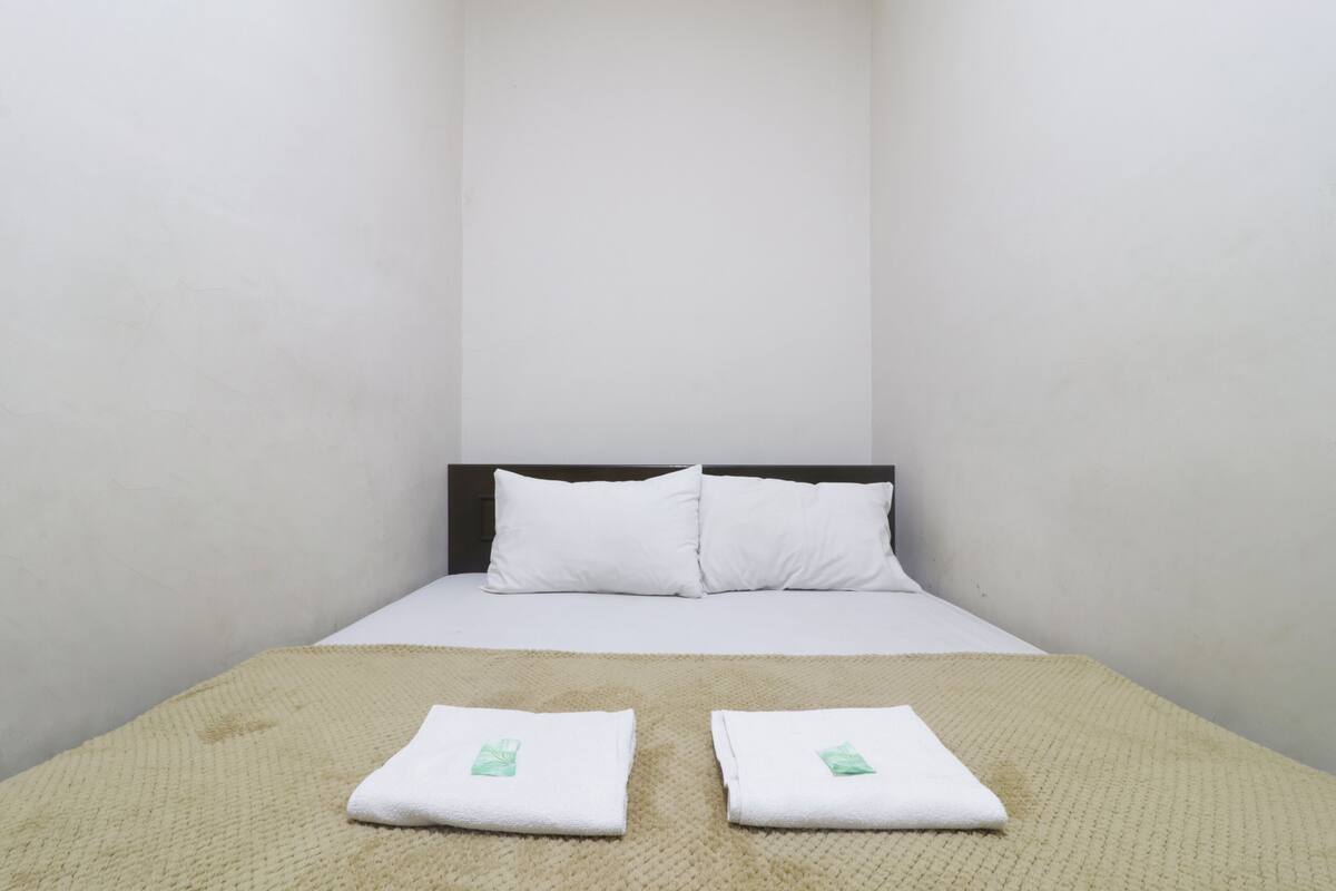 A bed with white linens, two pillows, and two folded towels with a green logo on a textured beige blanket.