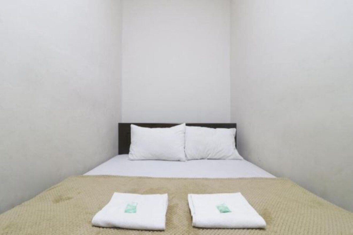 Una habitación de hotel sencilla y sin adornos, con una cama individual, ropa de cama blanca y una manta color beige.