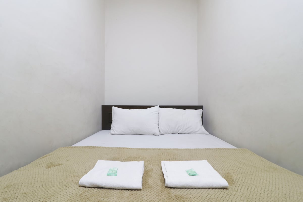 A single bed with white bedding and two folded towels with a green logo on a beige textured blanket.