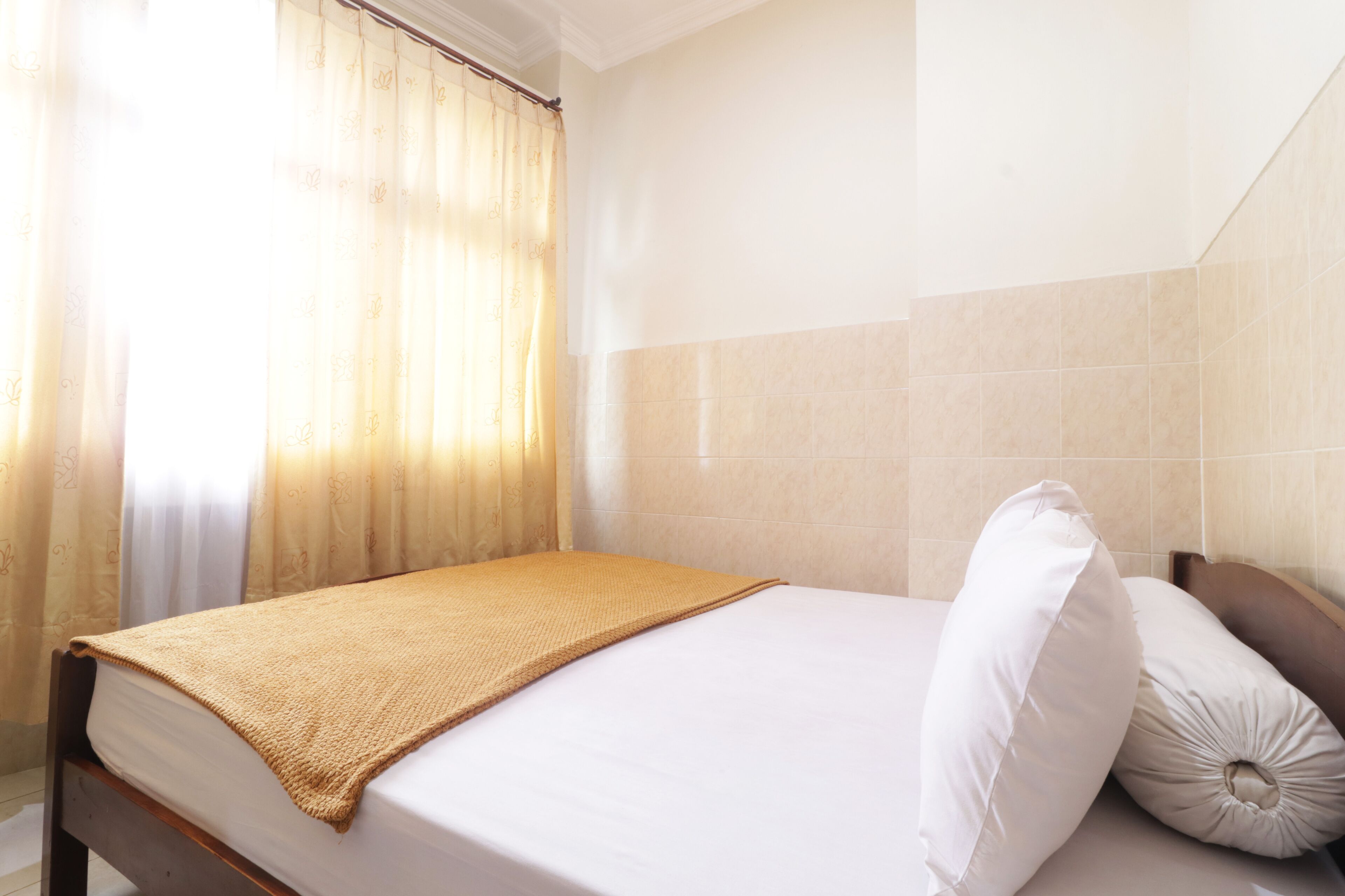 A neatly made bed with a beige blanket and white pillows, a wooden headboard, and a window with sheer curtains.