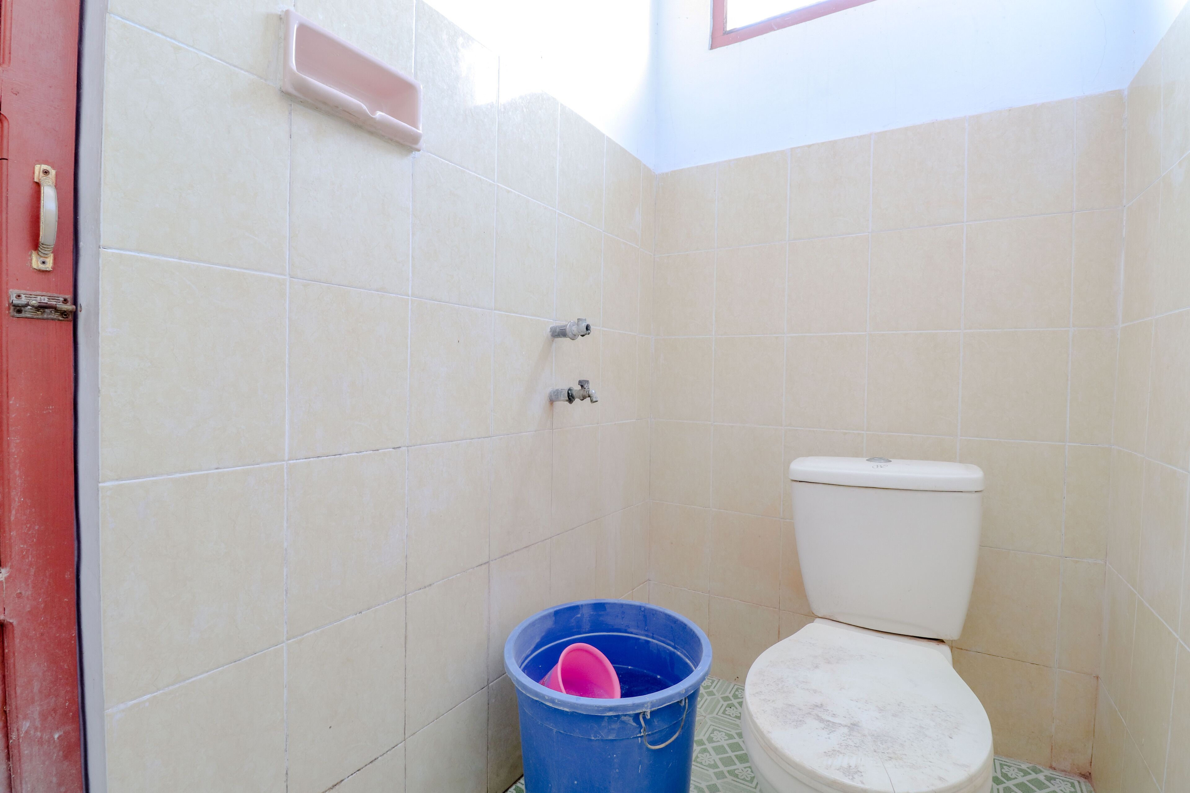 A small bathroom with a toilet, a blue bucket, and a red door.