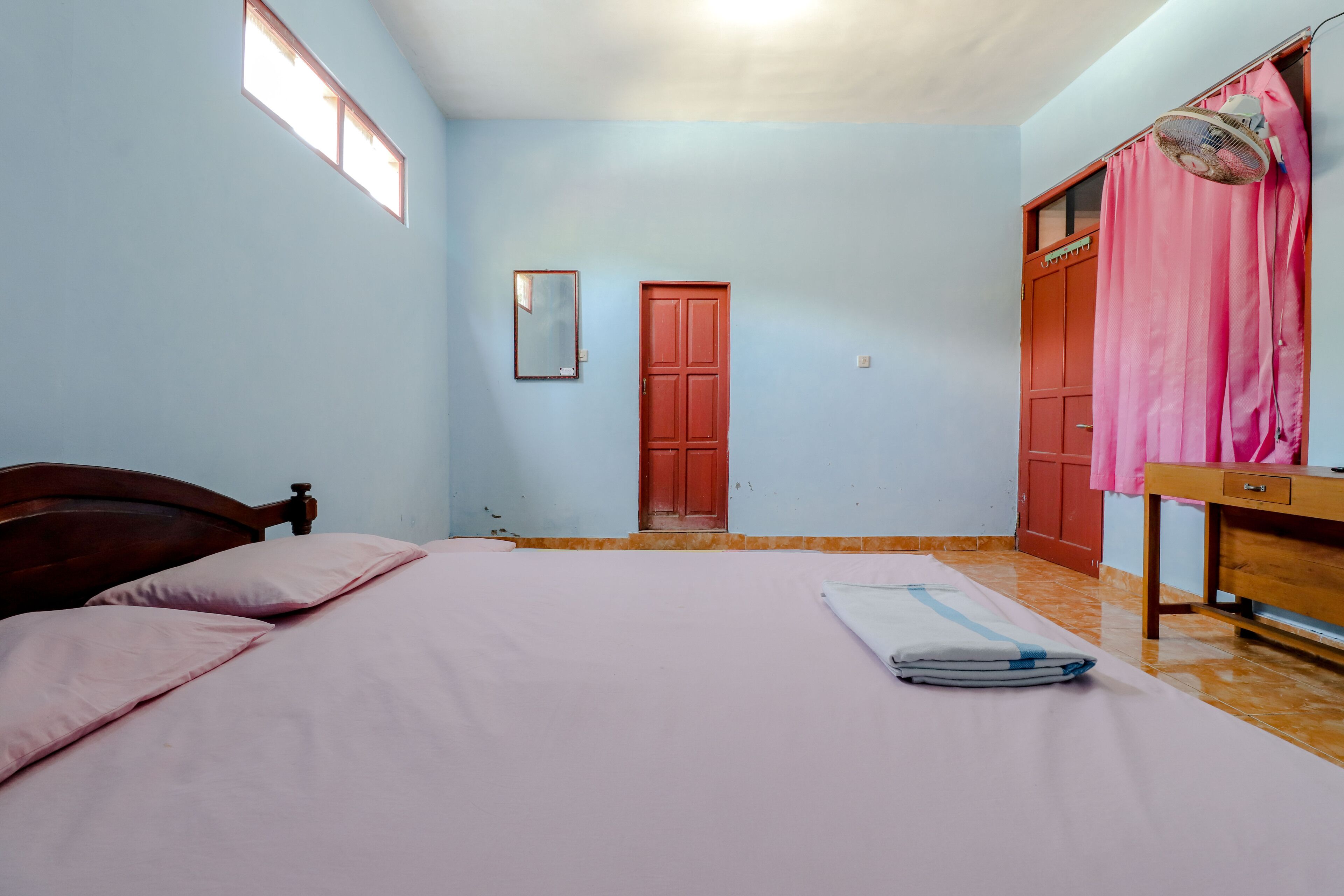 A bedroom with a bed, a wooden headboard, a red door, a window, and a fan.