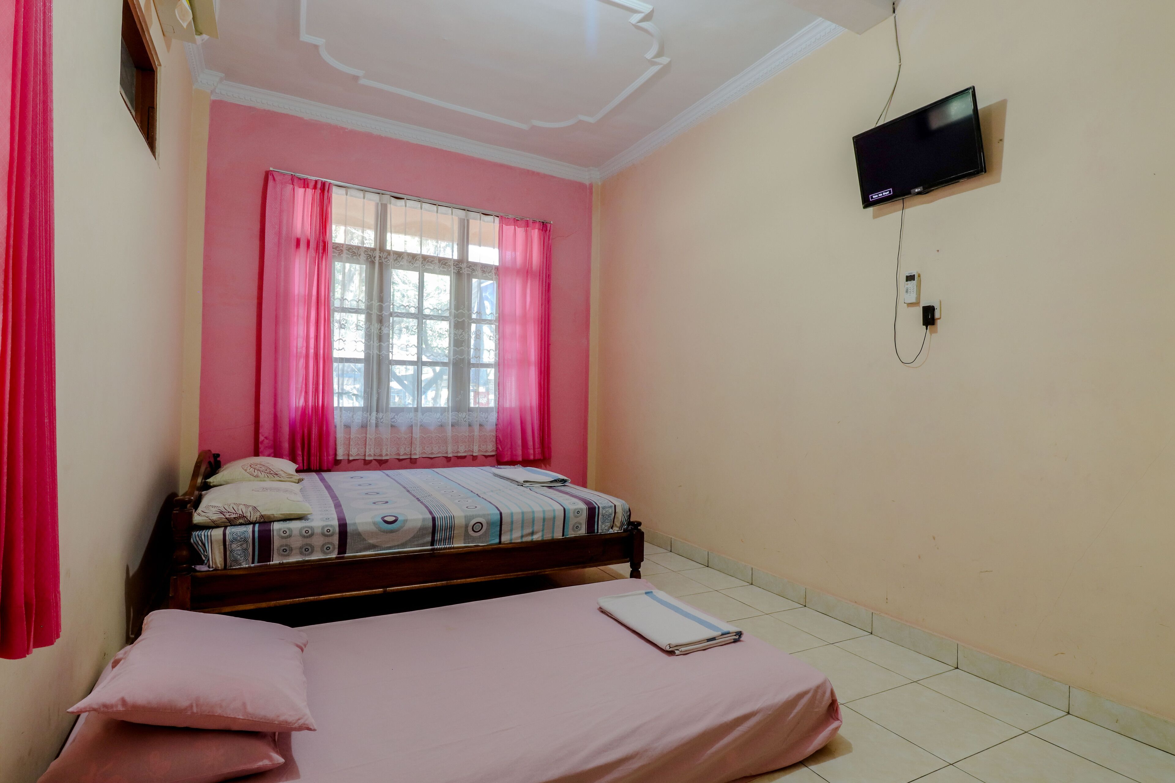 Un dormitorio con una cama, un televisor montado en la pared y una ventana con cortinas rosadas.