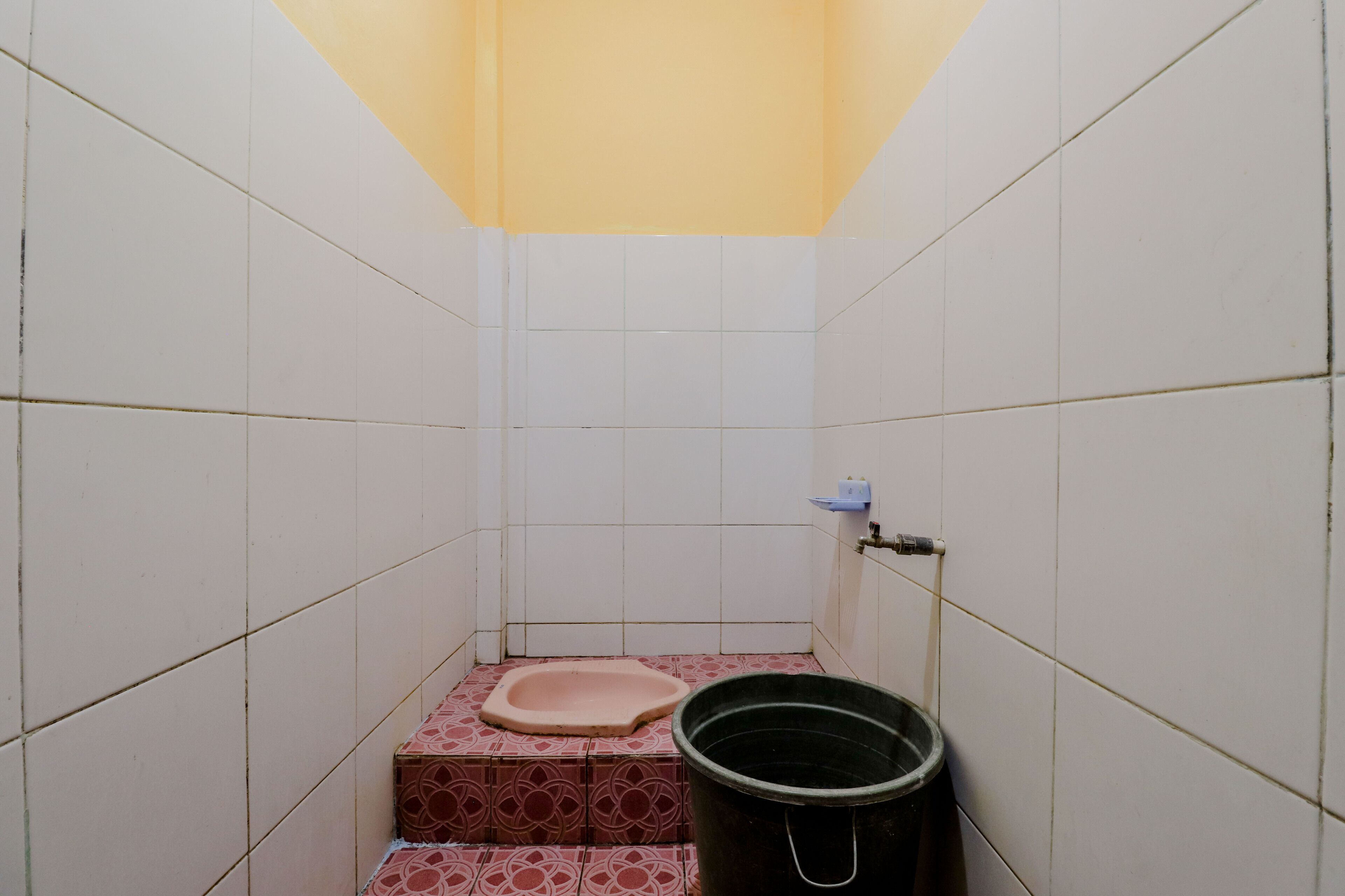 Un baño pequeño con azulejos, un inodoro, un balde y una repisa con un recipiente.