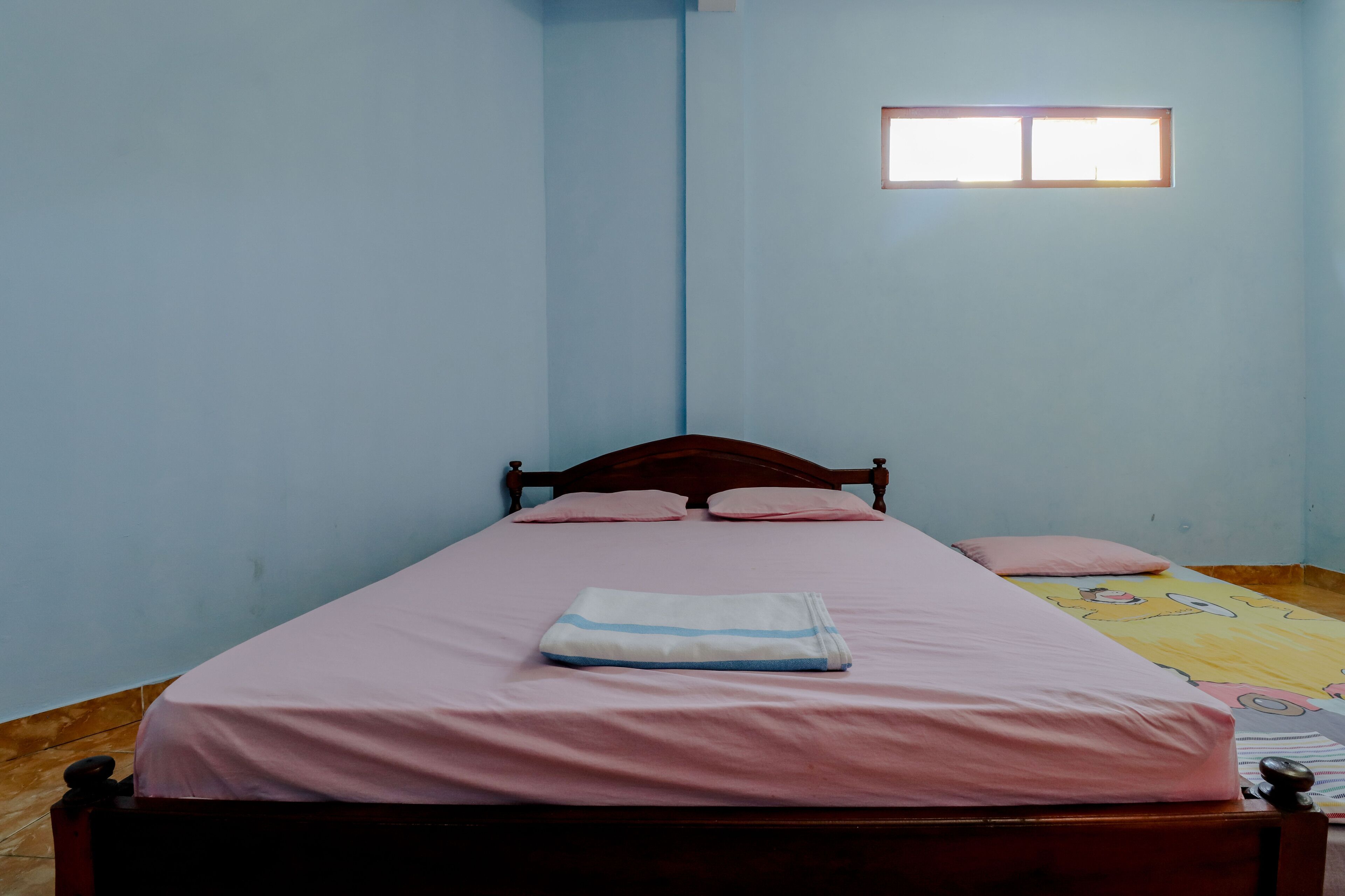 A single bed with a pink bedspread and pillows, a small window with natural light, and a wooden headboard.