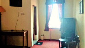 A room with a television mounted on the wall, a desk with a lamp, a chair, and a window with curtains. - Wisma & Kantin Amanda (Pacet)