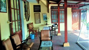 A room with a wooden bench, a table with a patterned tablecloth, and a chair. There is a window with a view of greenery outside. - Wisma & Kantin Amanda (Pacet)
