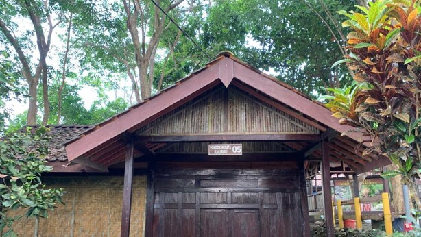 Un edificio de madera rústico con una puerta grande, dos sillas de madera y una motocicleta estacionada adelante.