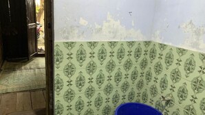 Un baño con baldosas verdes estampadas, un cubo azul y una puerta de madera que da a otra habitación.