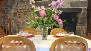 A dining table set with wicker chairs, a vase with flowers, and a stone wall in the background. - Chaplain's House: Charm in Swiss Normandy, Pets Allowed (Rabodanges)