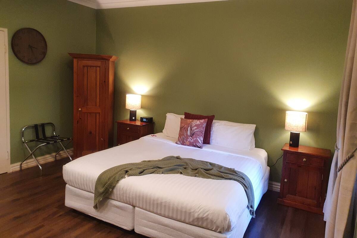 A bedroom with a bed, two bedside tables with lamps, a wooden wardrobe, a chair, and a clock on the wall.