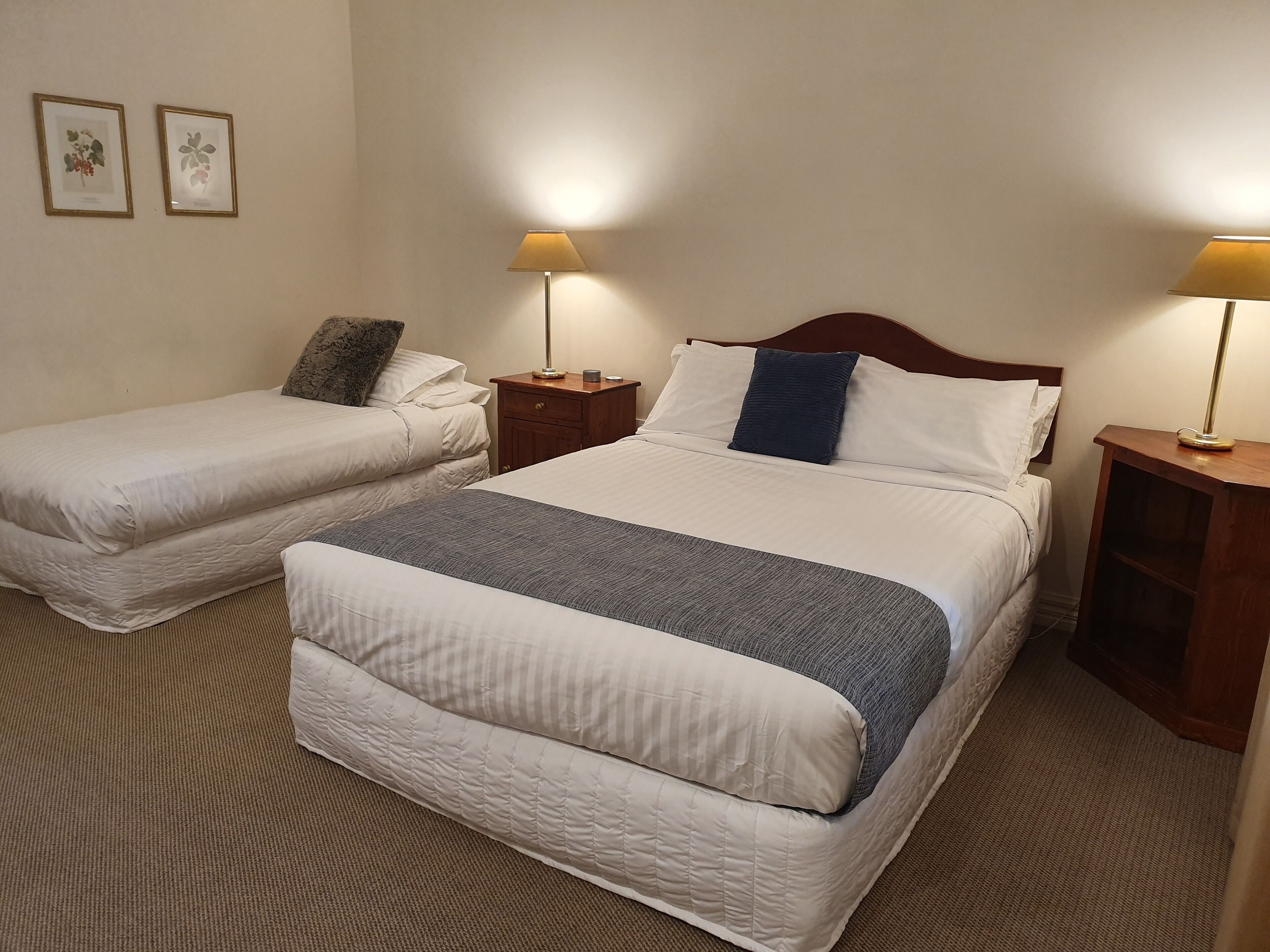 A hotel room with two single beds, a large bed, a nightstand with a lamp, and a small wooden shelf.