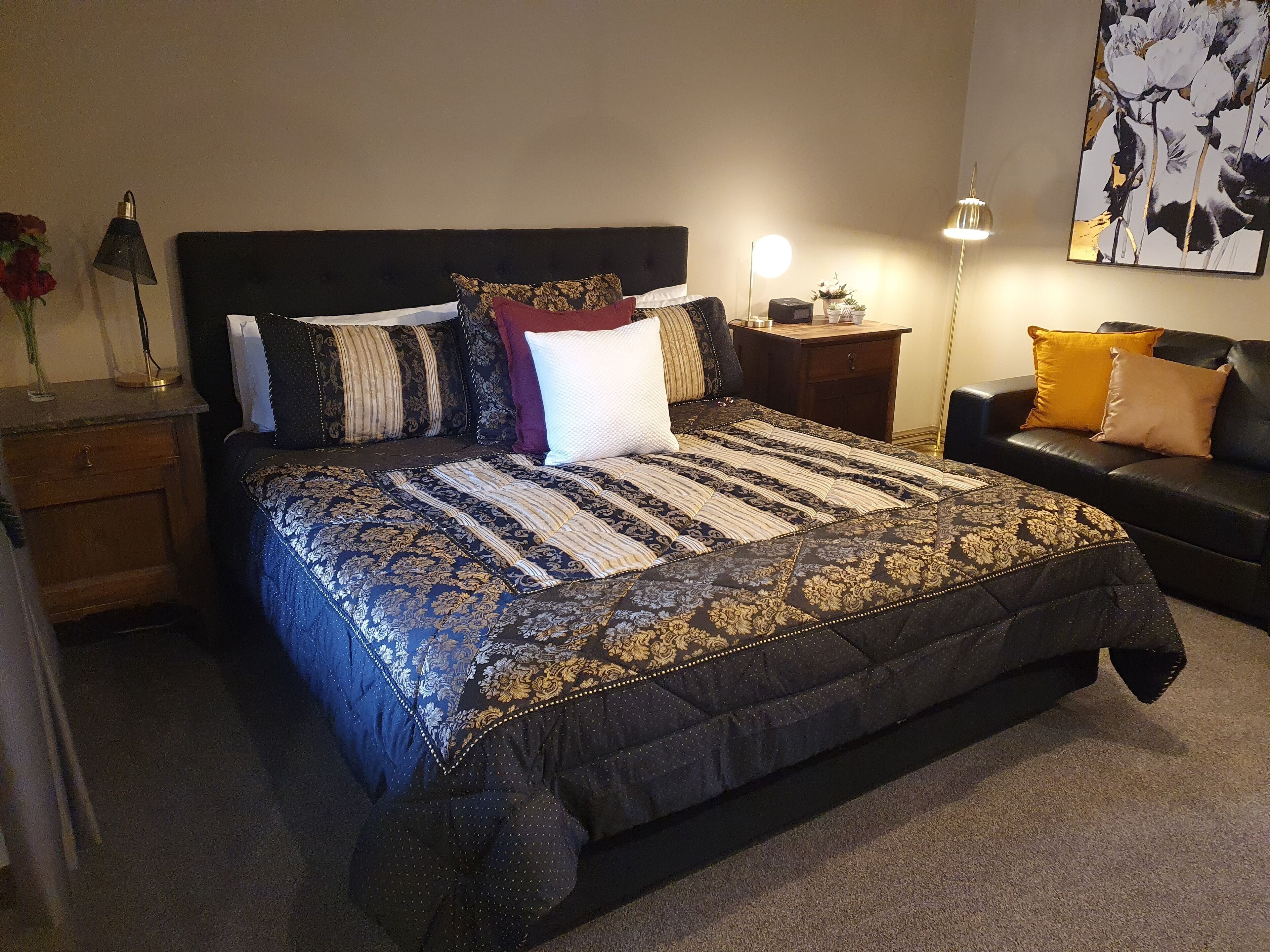 A bedroom with a large bed, a nightstand, a bedside table, a lamp, a couch, and a wall with a floral painting.
