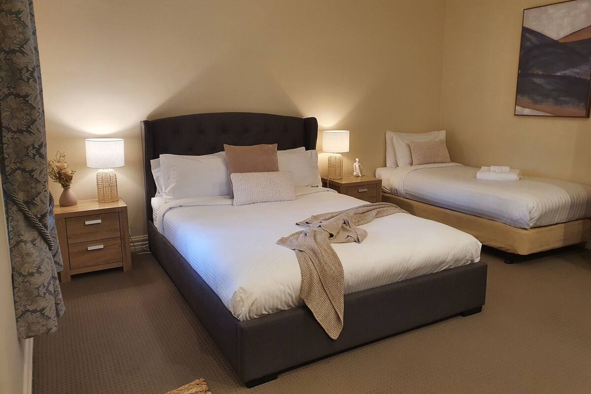 A hotel room with a large bed, a smaller bed, two bedside tables with lamps, a nightstand, and a painting on the wall.