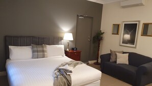 A hotel room with a large bed, a bedside table with a lamp, a dark-colored sofa, and a door. - The Lancefield Lodge (Lancefield)