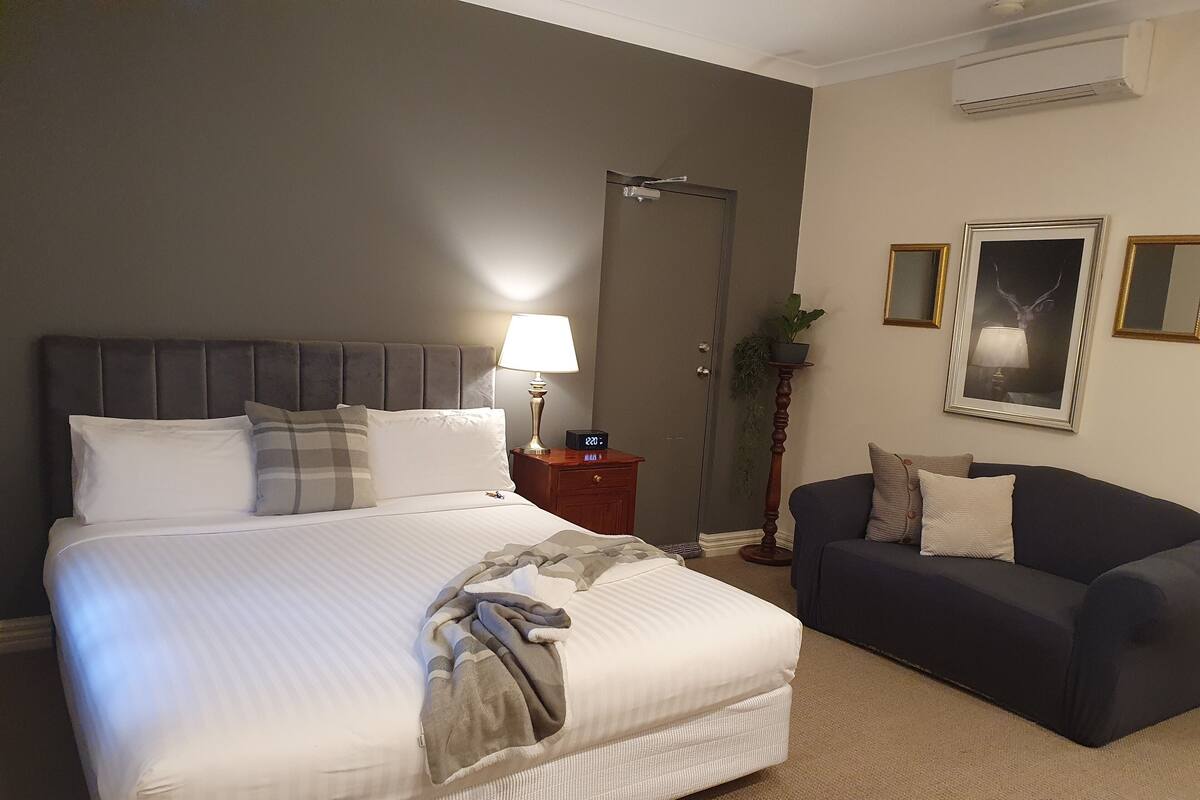 A hotel room with a large bed, a bedside table with a lamp, a dark-colored sofa, and a door.