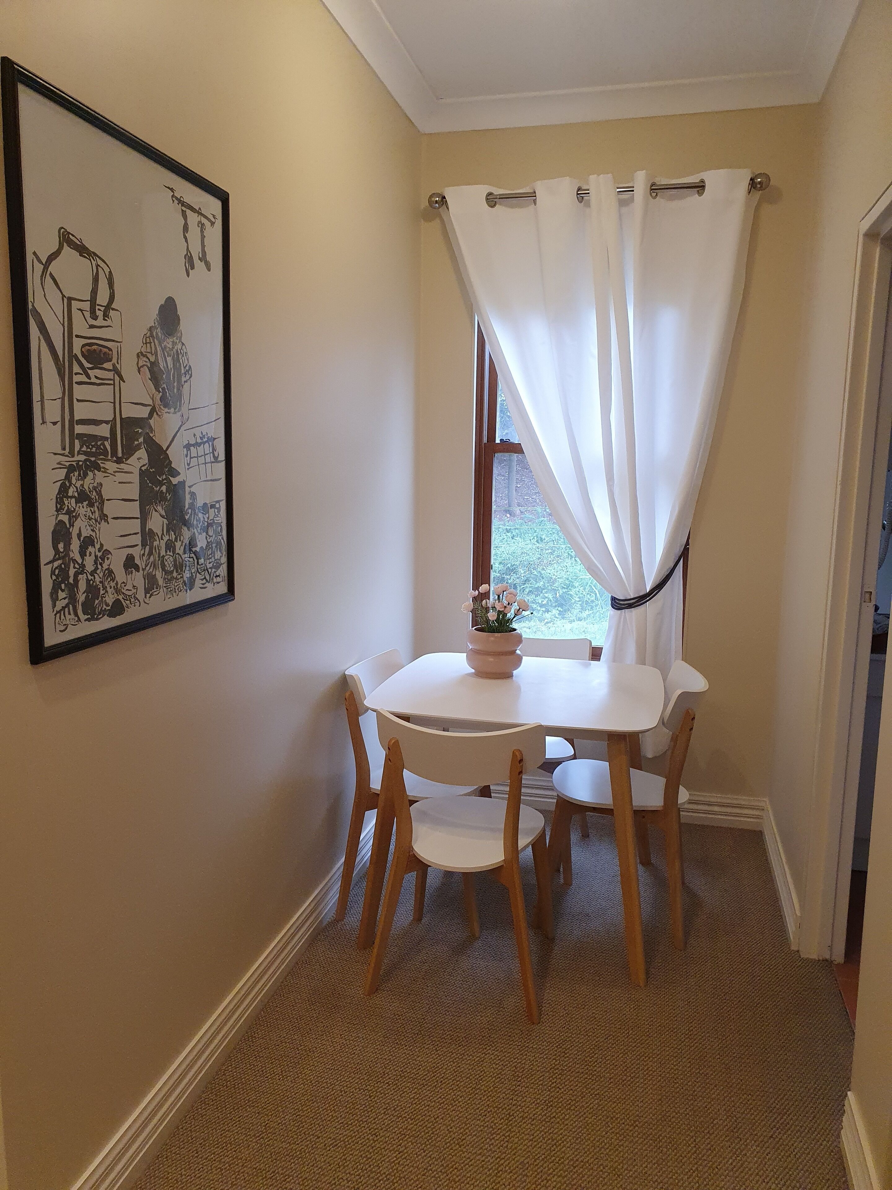 A dining area with a white table and chairs, a framed artwork on the wall, and a window with white curtains.