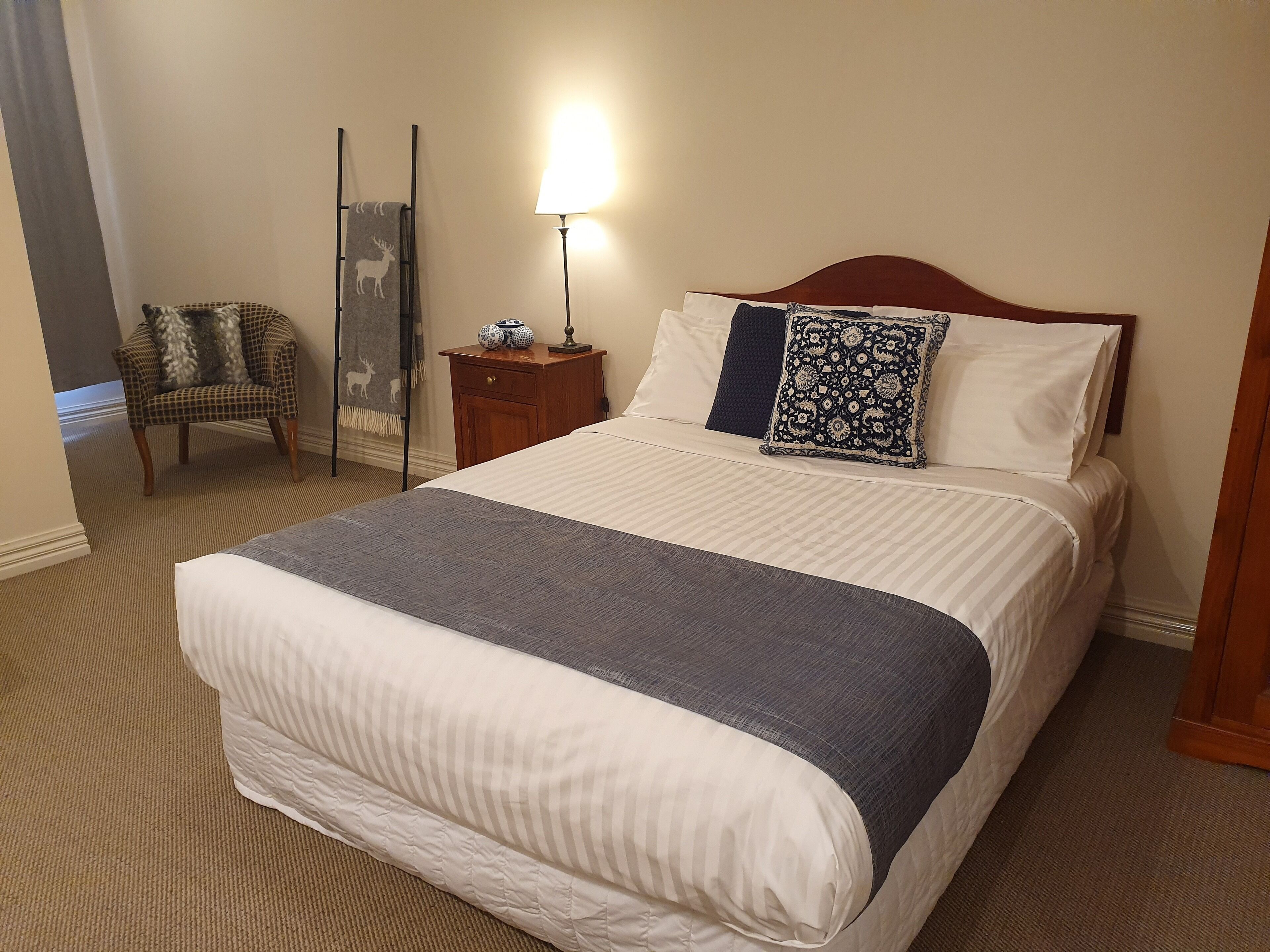 Una cama bien tendida con una manta gris, una mesita de noche de madera con una lámpara, una silla con un cojín estampado y un espejo decorativo.