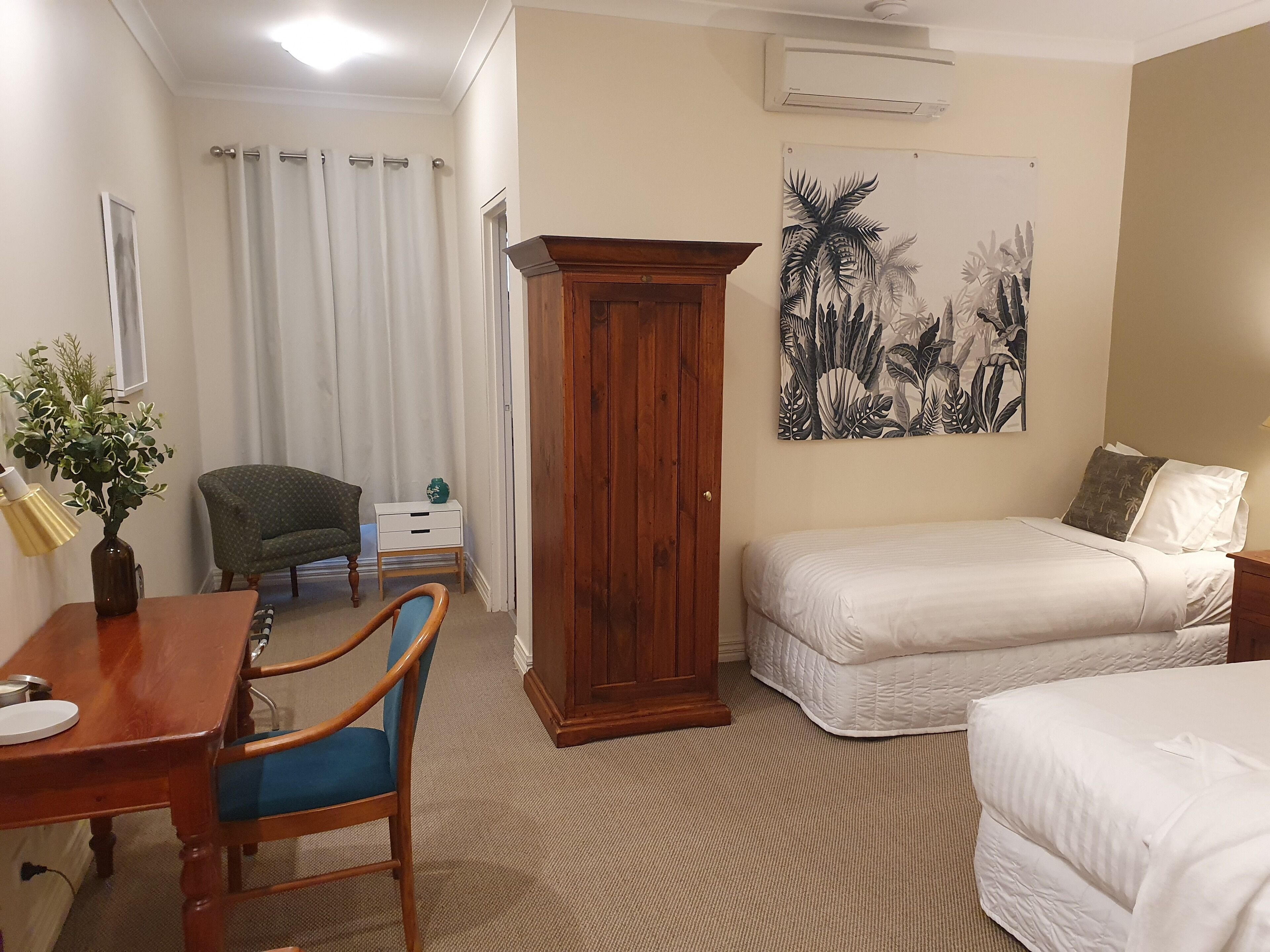 Une chambre d’hôtel avec un bureau en bois, une chaise, un lit avec une literie blanche, une table de chevet, une armoire, un climatiseur mural et un tableau sur le thème des pays tropicaux.
