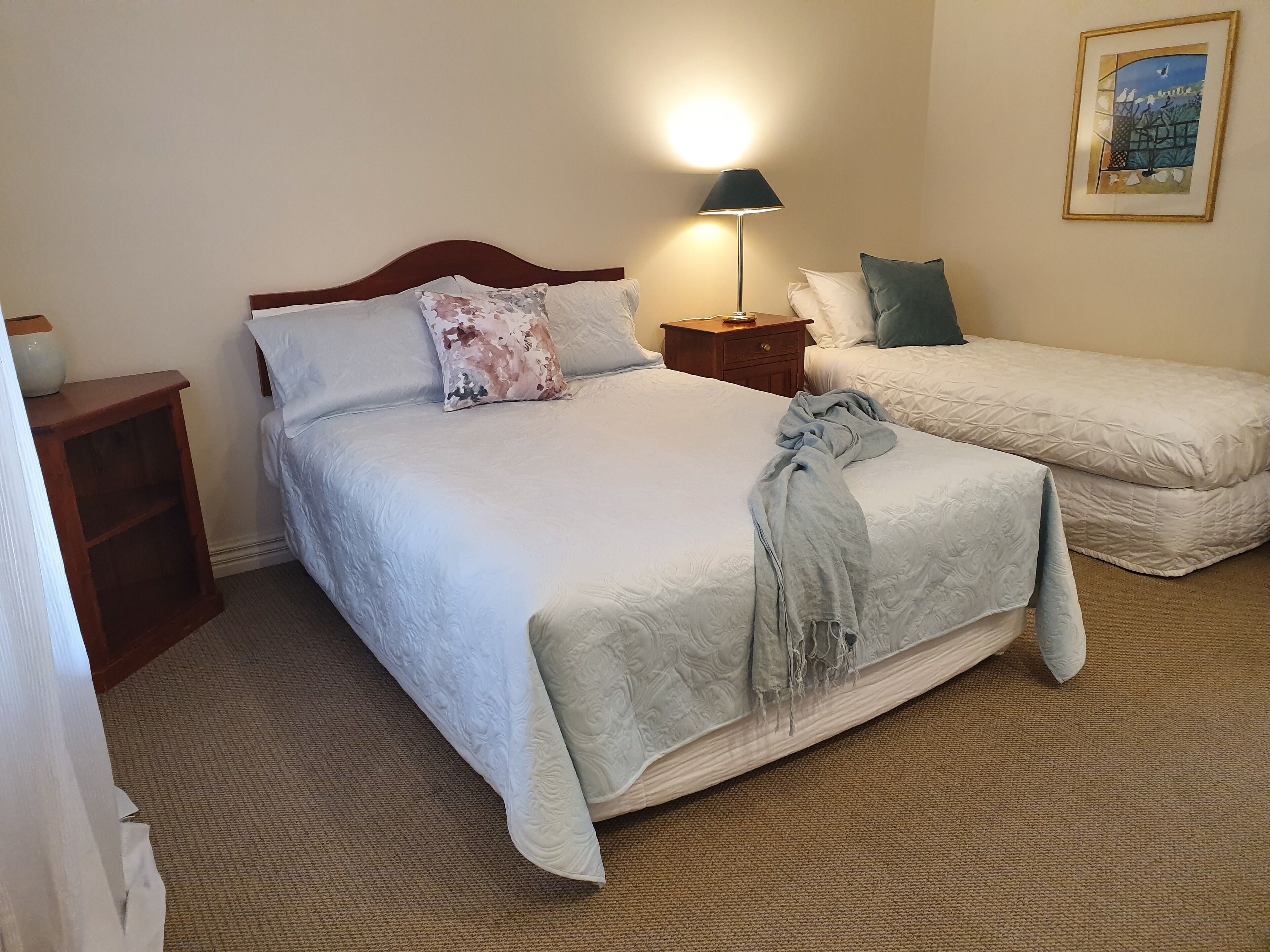 A hotel room with a large bed, a smaller bed, a nightstand with a lamp, a wooden side table, and a framed picture on the wall.