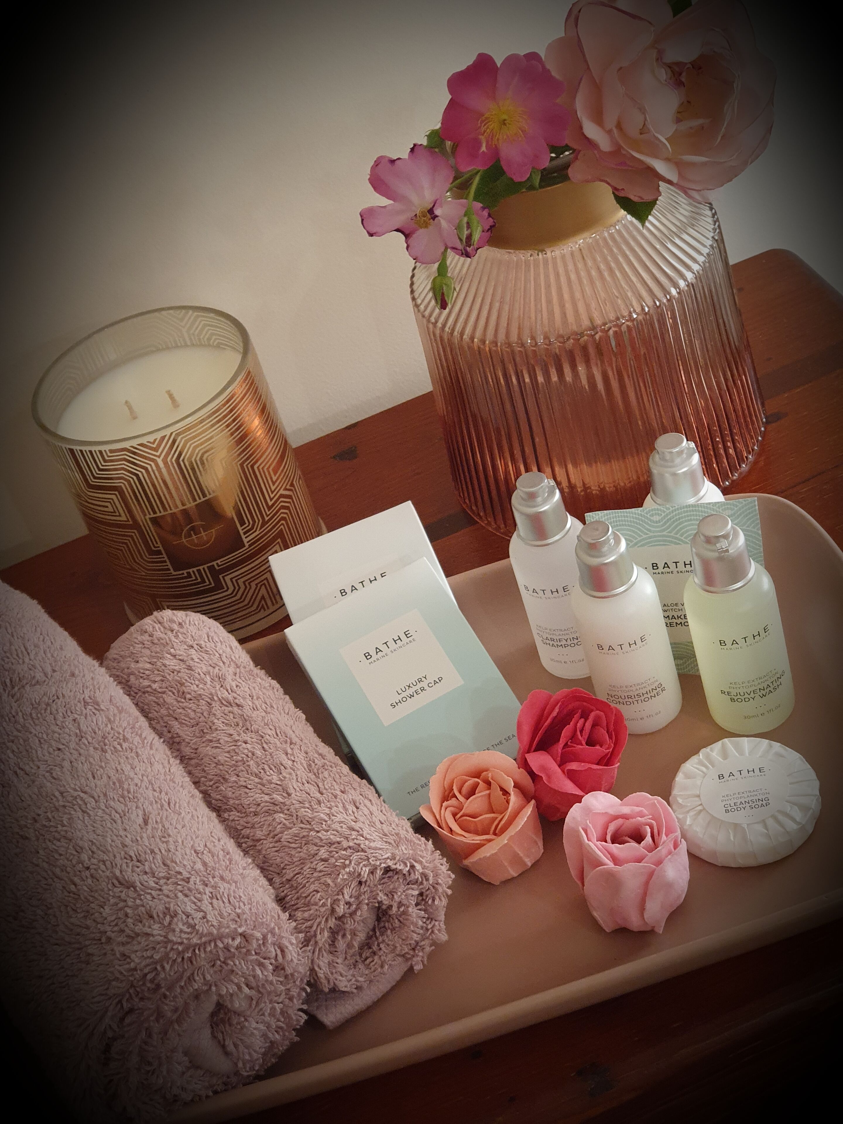 Ett bricka med toalettartiklar, inklusive ett lavendeldoftande ljus, en glasvas med rosa blommor och olika produkter från Bath & Body Works.
