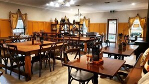 A dining area with multiple wooden tables and chairs, a chandelier, and a bookshelf with decorative items. - The Lancefield Lodge (Lancefield)
