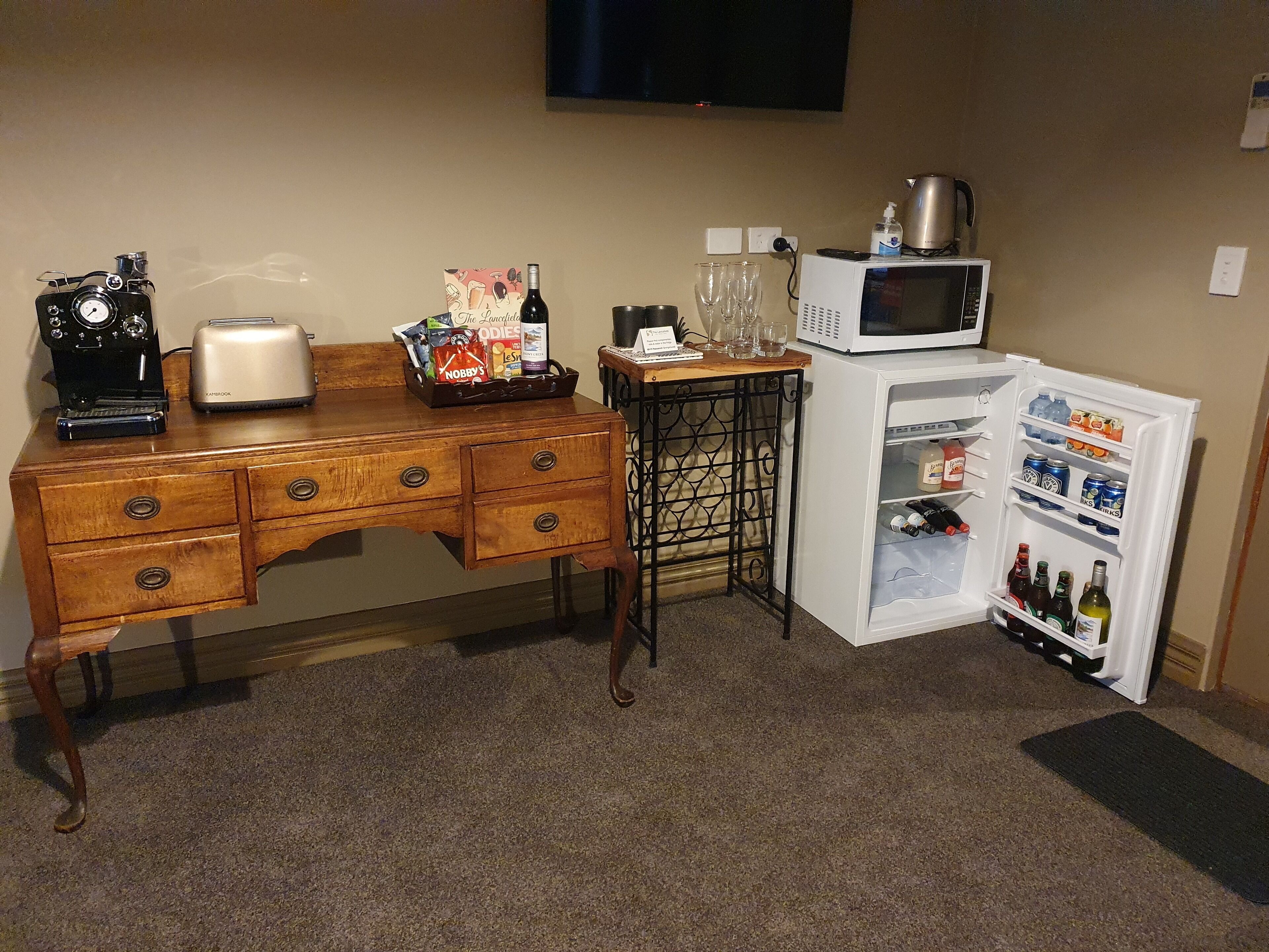 A wooden desk with a vintage camera, a toaster, and a tray of snacks. A small table with a kettle and glasses is next to a white refrigerator stocked with beverages.
