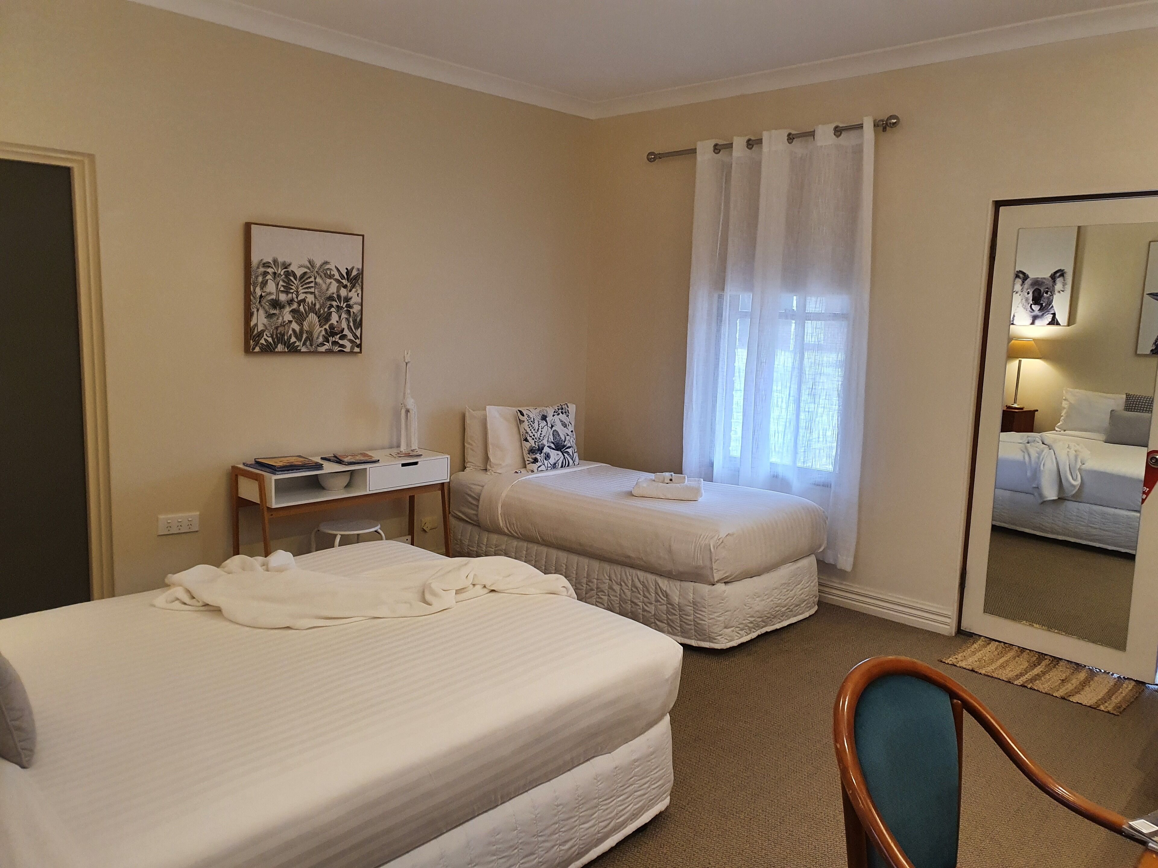 A hotel room with two beds, a desk with a chair, a window with curtains, and a mirror on the wall.