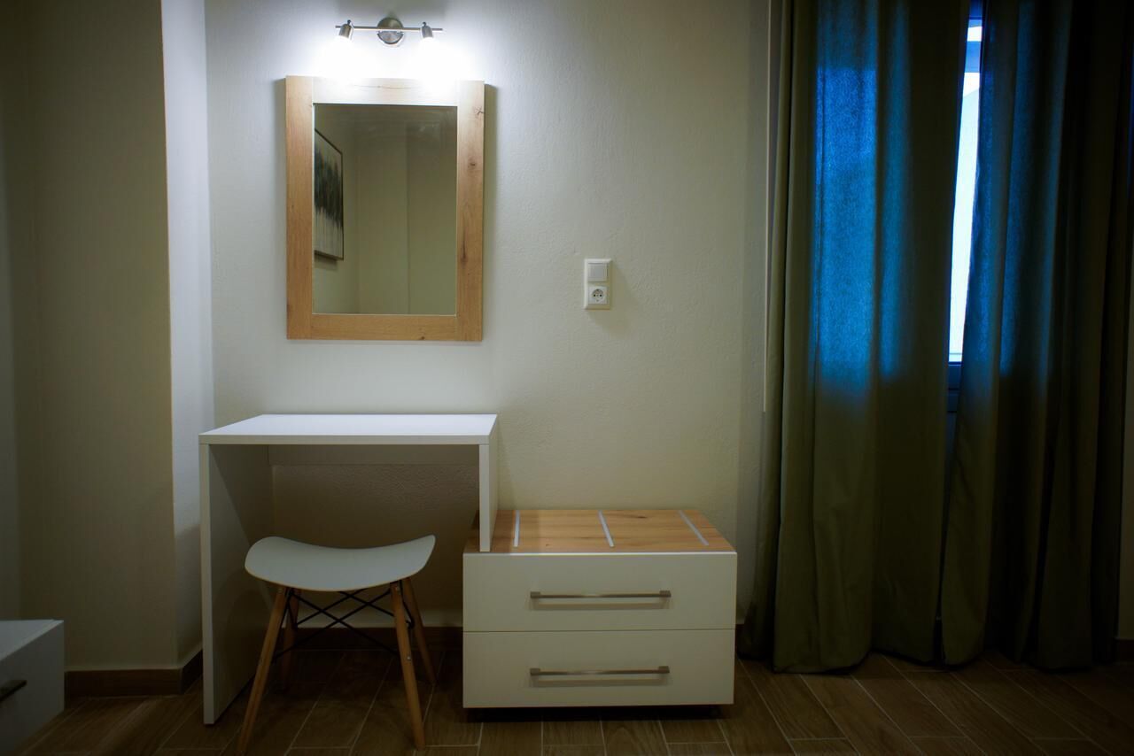 Een kleine, privé badkamer met een wit bureau, een houten krukje, een spiegel en een klein houten kastje met lades.